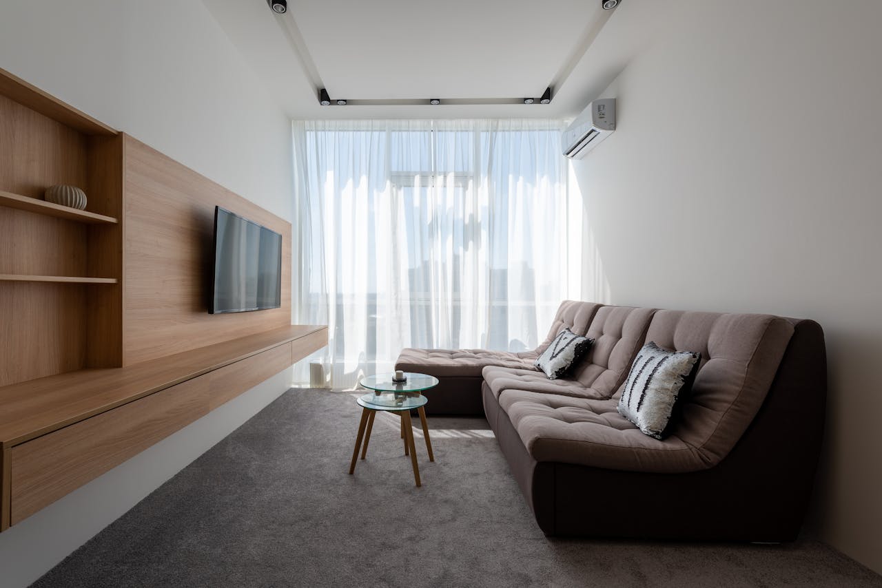 Comfortable couch with cushions near side tables against wall with wooden cupboard and modern TV in stylish living room with window